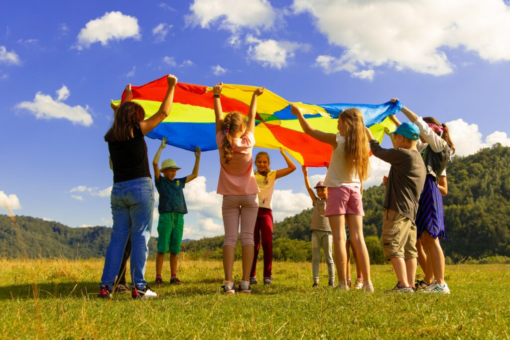 Eleven children (about 11 years old) hold up a multicoloured parachute (red, yellow, blue, and green). The children are smiling and having fun with their parachute game in the field with forested mountains int he background and a bright blue sky.